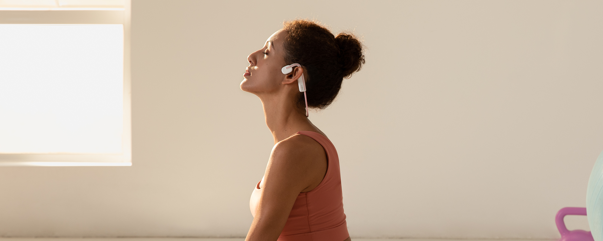 Image of a woman in an empty, well-lit room with eyes closed wearing Shokz OpenMove headphones