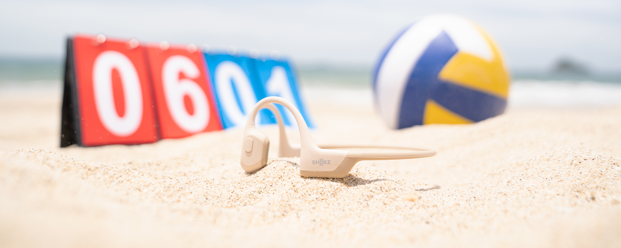 Image of Beige OpenRun Pro wireless headphones in the sand next to a volleyball and scoring card