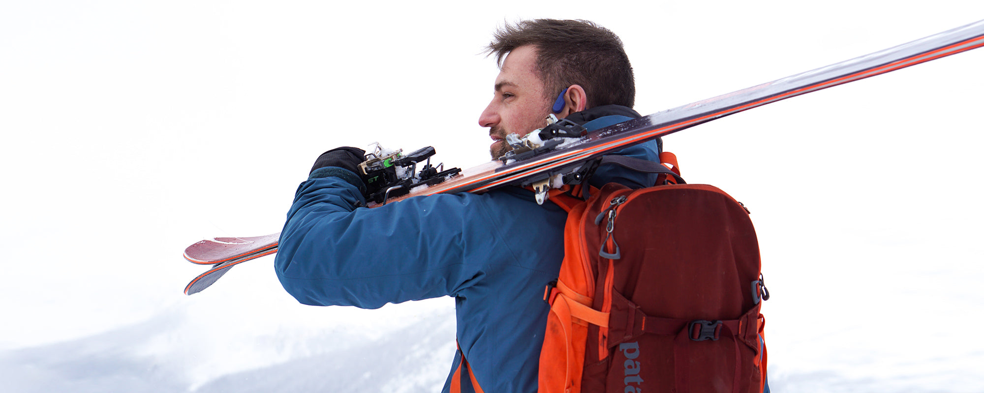 Man holding skiis on snowy mountain and wearing AfterShokz Aeropex headphones